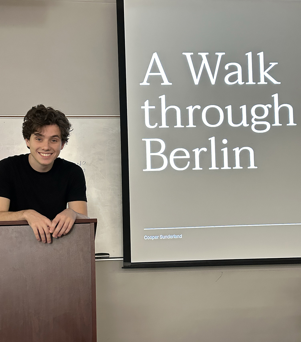 photo of man at lectern, screen with text in background