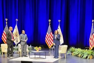 photo of three people on stage with flags, chairs, table