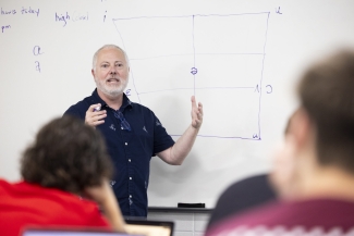 photo of man gesturing, whiteboard in background, students in foreground